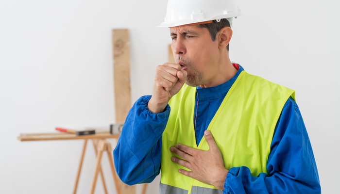 A male construction worker in a hard hat and a high-visibility vest coughing into his fist with his hand on his chest.