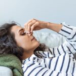 A woman wearing a striped shirt is lying on an arm rest of a couch. She is holding the bridge of her nose and closing her eyes.