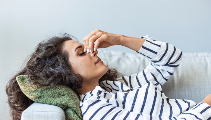 A woman wearing a striped shirt is lying on an arm rest of a couch. She is holding the bridge of her nose and closing her eyes.