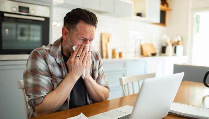 A man sits at a kitchen table and sneezes into a tissue while using a laptop, illustrating indoor allergies and poor air quality.